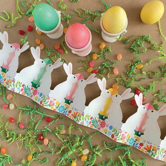 Top-down flatlay of printable standing bunny garland with Easter grass, dyed eggs, egg cups, and jelly beans – accordion fold tabletop centerpiece digital download