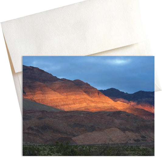 A photo titled "Winter Warmth" captures the contrasting elements of a Death Valley winter sunset.  A rugged mountain range dominates the background, its face highlighted by a band of golden sunlight that cuts through the cloudy sky.