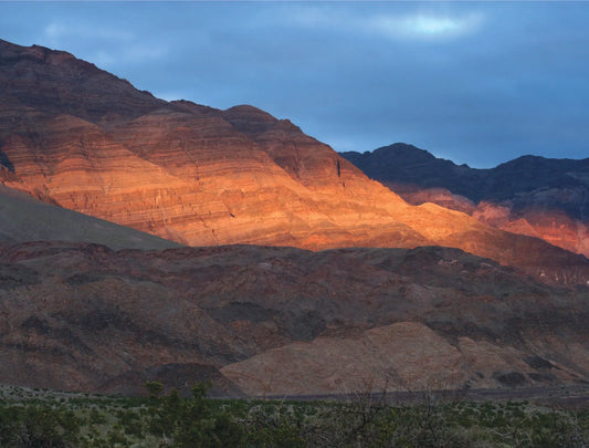 A photo titled "Winter Warmth" captures the contrasting elements of a Death Valley winter sunset. A rugged mountain range dominates the background, its face highlighted by a band of golden sunlight that cuts through the cloudy sky.