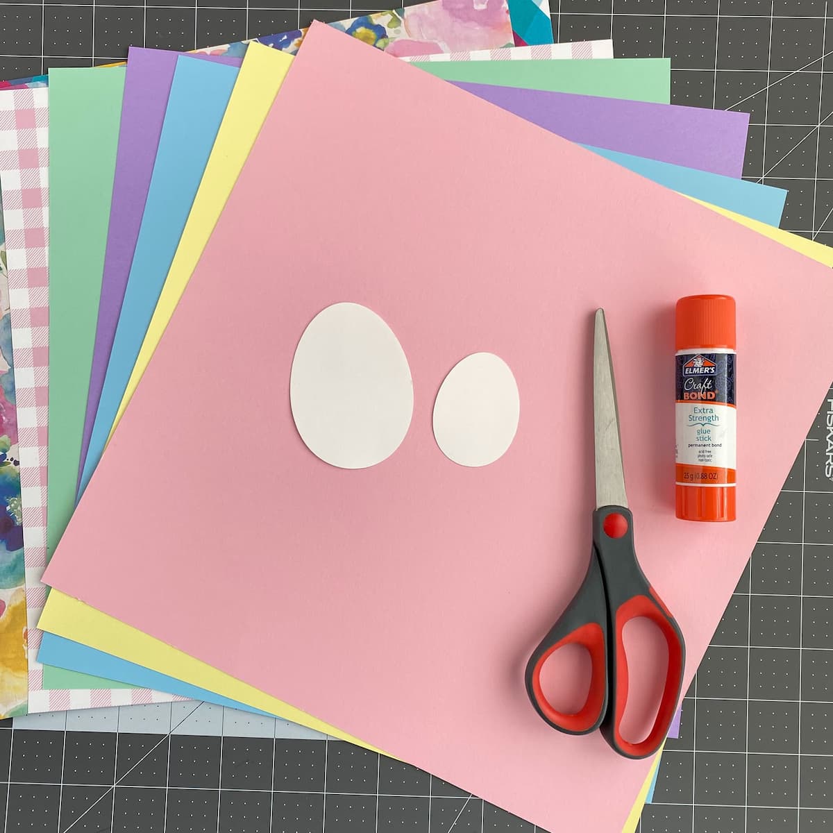 Colorful paper sheets with cut-out egg shapes, scissors, and glue on a grid background