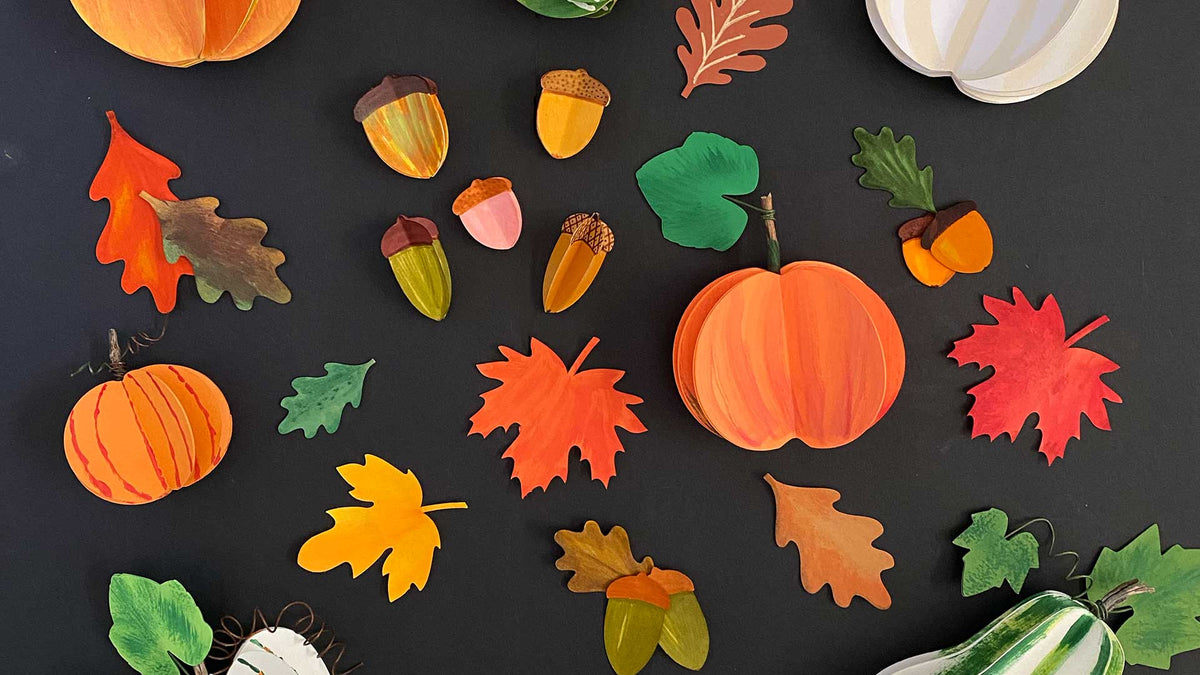 A flat-lay arrangement of autumn-themed paper crafts and decorations on a dark background, including orange pumpkins, colorful fall leaves, acorns, and small gourds.