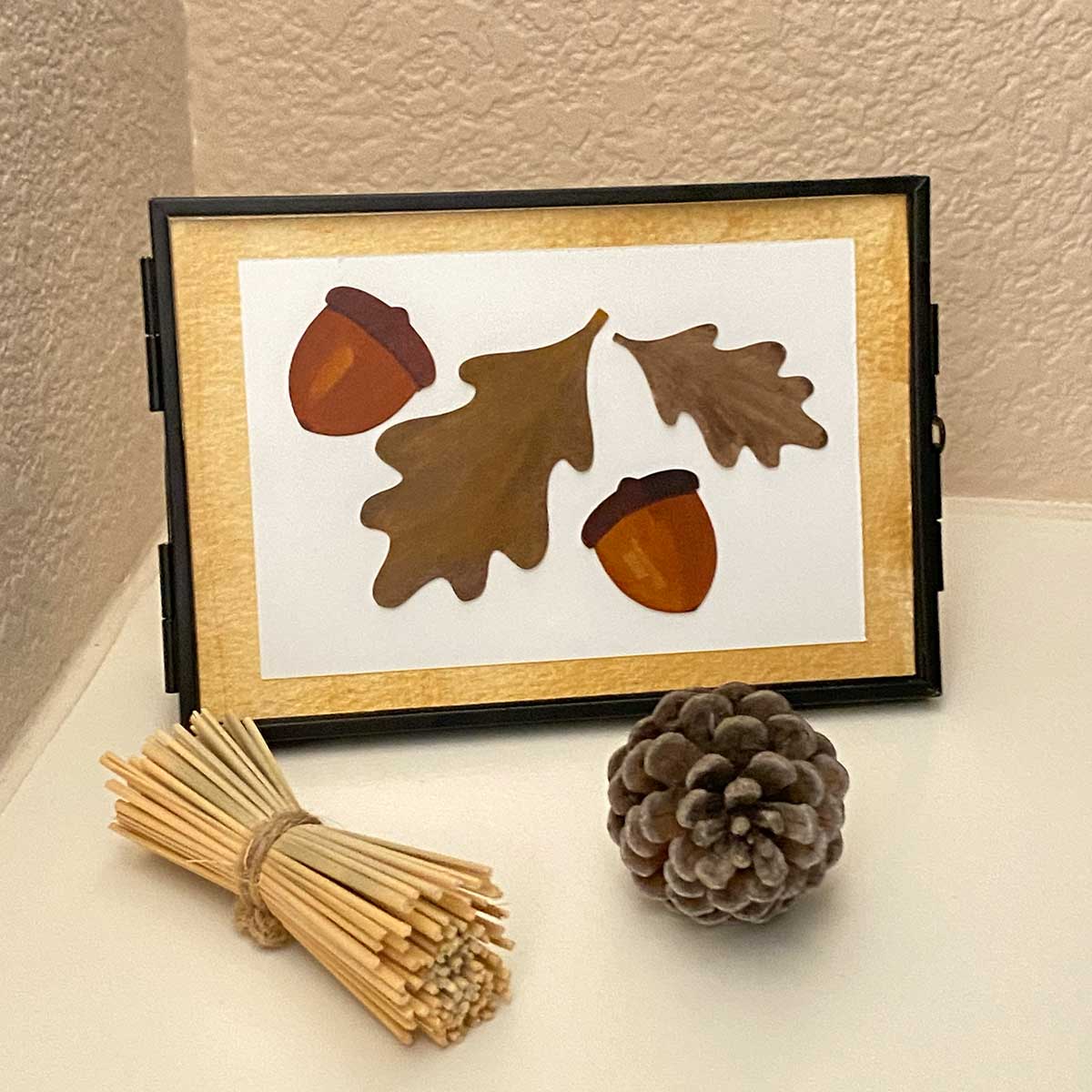 Framed artwork with acorns and leaves on a white surface with a pine cone and sticks for CakewalkDIY Paper Craft tutorial.