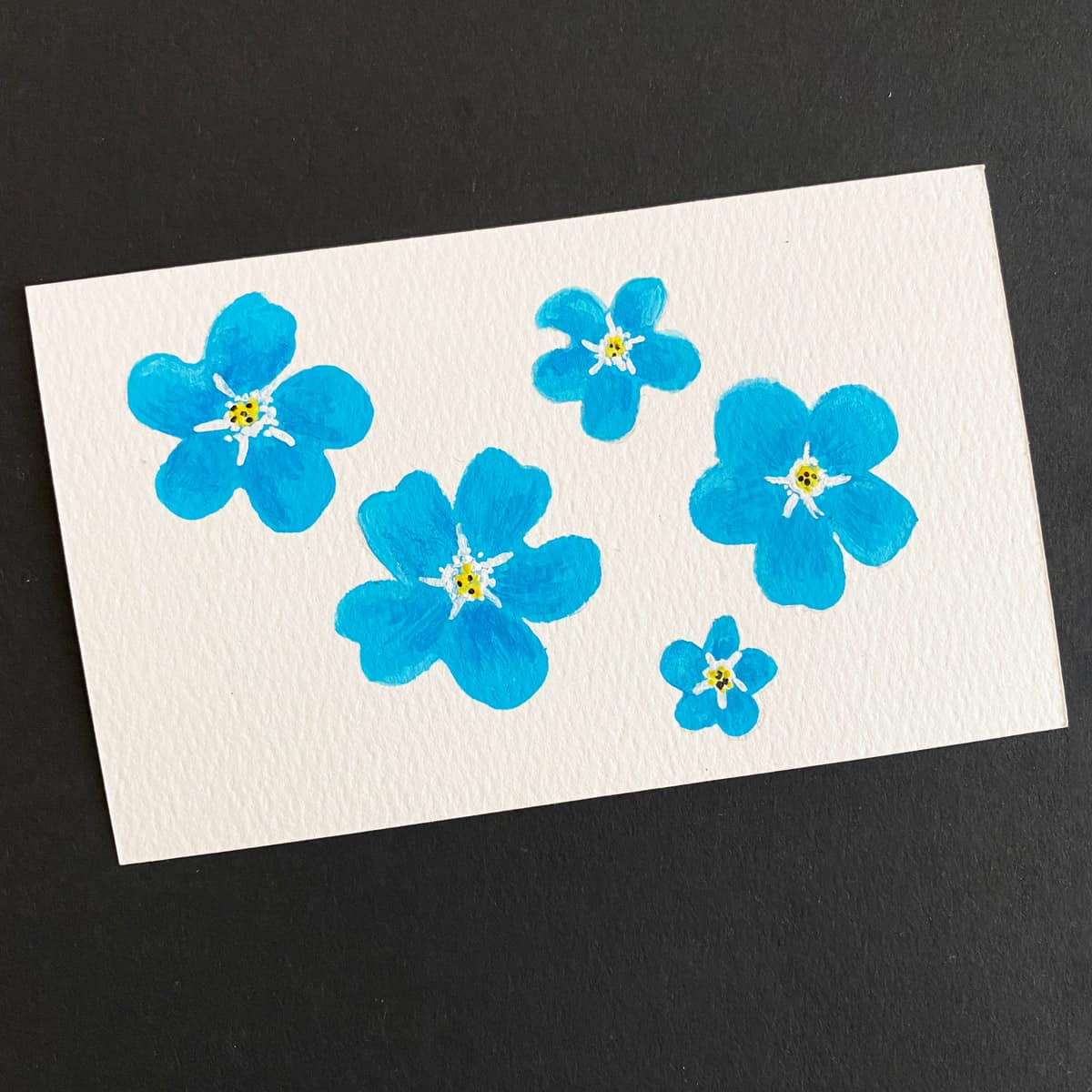 Index card with a painting of forget-me-not flowers made with gouache watercolors on a black background by Mozaic Studio for ICAD 2025
