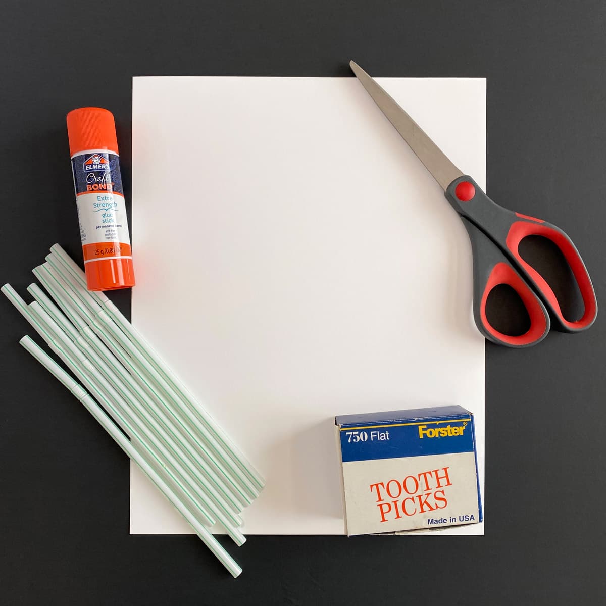 Supplies including glue, scissors, straws, and toothpicks on a white sheet of paper with a black background.